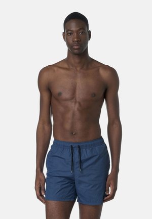 Giovane uomo in piedi di fronte, indossa pantaloncini da nuoto blu con coulisse nere, su sfondo grigio chiaro uniforme.