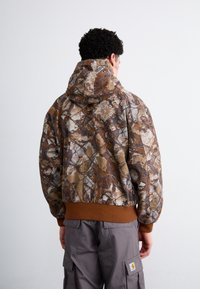 Camo-gepatroneerde hoodie met een bruin en tan kleurenschema, voorzien van een trekkoordkap en een geribde bruine onderrand. Zacht materiaal en een ontspannen pasvorm.