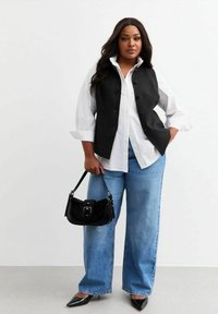 Chaleco negro superpuesto sobre una camisa blanca de botones, combinado con pantalones de mezclilla anchos en un tono azul claro y un bolso negro con detalle de hebilla. Zapatos negros de punta.