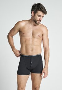 Black cotton boxer shorts with a buttoned fly, elastic waistband featuring a logo, and a snug fit. Worn by a male model.