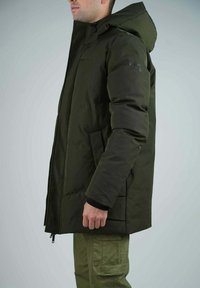 Veste parka rembourrée en vert olive foncé avec capuche, fermeture éclair à l'avant et poches latérales. Présente une texture lisse et mate.