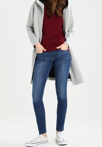Felpa grigia lunga con una maglietta bordeaux sotto, abbinata a jeans blu aderenti e sneakers bianche. L'outfit presenta un design casual.