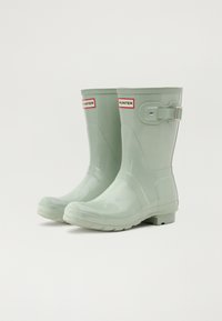 Bottes de pluie en caoutchouc vert avec une finition glossy, un détail de boucle sur le côté et une semelle robuste. Présente le logo "HUNTER" sur des accents rouges.