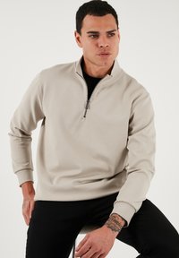 Beige pulóver so štvrťzipsom z textúrovaného materiálu s dlhými rukávmi a rebrovanými manžetami. Model je posadený a má na sebe čierne oblečenie pod ním.