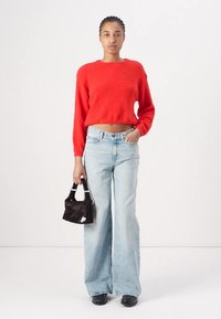 Pull court rouge à finition texturée, associé à un jean large délavé. Le mannequin tient un petit sac à main noir. Des chaussures noires complètent le look.