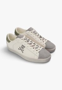 Zapatillas de piel blanca con acentos de ante gris en la puntera y el talón. Presentan un logo de calavera y tibias grises, diseño de cordones y suela texturizada.