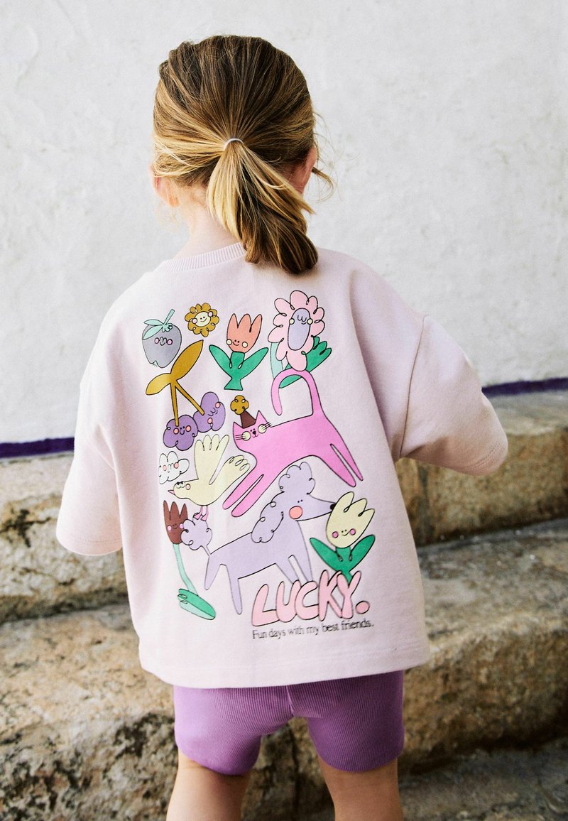 Enfant aux cheveux blonds en queue de cheval portant un t-shirt rose avec des animaux de dessin animé colorés et des fleurs, debout sur des marches en pierre, de dos.