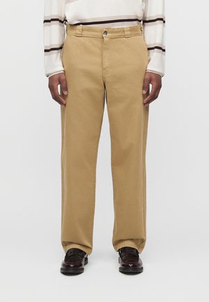 Homme portant un pantalon beige à jambes droites, des mocassins marron foncé et une chemise blanche à manches longues avec des rayures horizontales noires et marron.
