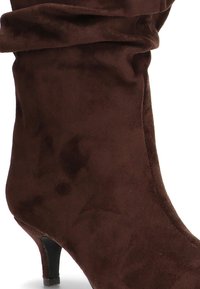 Botín de ante marrón con tacón cónico, diseño holgado y textura suave. Presenta un puño plegado en la parte superior.