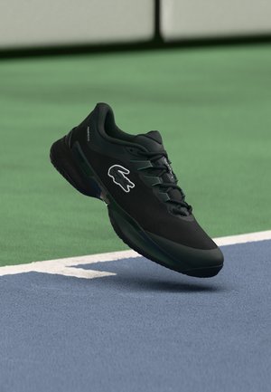 Chaussure de sport noire Lacoste avec lacets flottant au-dessus de la ligne de limite d'un court de tennis sur une surface verte et bleue.