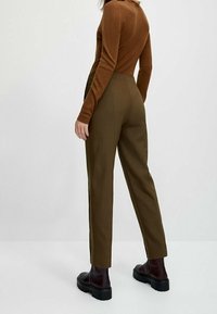 Brown fitted sweater with long sleeves paired with olive green tailored trousers and black chunky-sole ankle boots, viewed from behind.