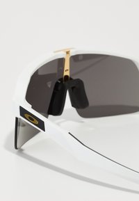 Gafas de sol blancas con grandes lentes grises, acentos de metal dorado y un panel lateral negro texturizado. Presentan un diseño angular deportivo.