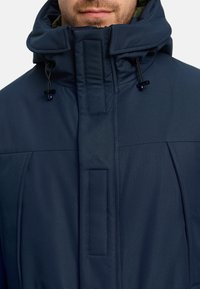 Marineblaue isolierte Jacke mit hohem Kragen, verstellbarer Kordel und dezentem Strukturmuster. Mit Reißverschluss und Knopfverschluss als Details.
