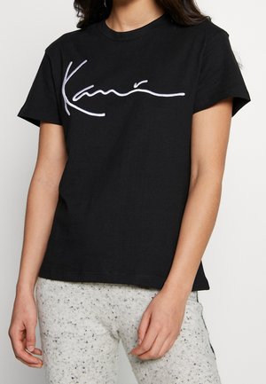 Kvinde iført en sort T-shirt med korte ærmer med hvid kursiv tekst og lysegrå, brogede bukser, stående mod en ensfarvet baggrund.