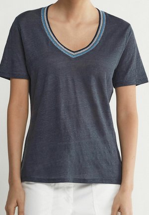 Donna che indossa una maglietta grigio scuro con scollo a V e maniche corte con bordi blu e bianchi, abbinata a pantaloni bianchi, mostrata dal collo ai fianchi.