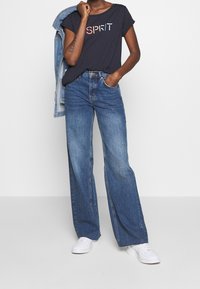 Camiseta navy con el logo "SPIRIT", jeans de pierna ancha azul y zapatillas blancas; el denim tiene una textura suave y un ajuste casual. Chaqueta de denim clara deslizándose por el brazo.