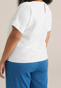 White short-sleeve top with a back keyhole detail, paired with blue denim pants. The fabric appears lightweight and textured.