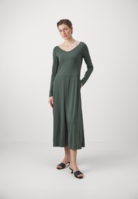 Robe à manches longues, vert foncé, avec un décolleté en V, une jupe fluide et des poches latérales, fabriquée en tissu doux ; assortie à des chaussures à talons ouverts noires.