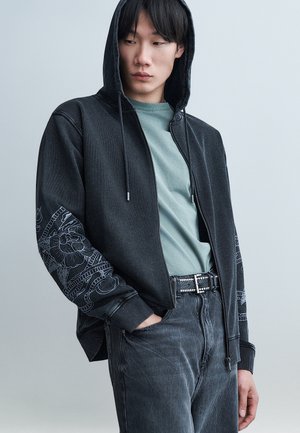 Jeune personne portant un sweat à capuche noir avec des motifs floraux sur les manches, un T-shirt vert menthe, une ceinture noire cloutée et un jean foncé, les mains dans les poches.