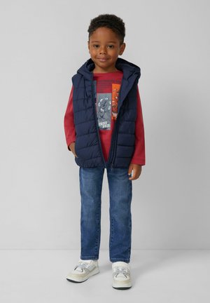 Ein kleiner Junge trägt eine navyblaue Pufferweste, ein rotes Langarmshirt mit Grafik, blaue Jeans und weiße Sneakers. Er steht mit den Händen in den Taschen.