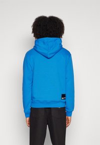 Blauwe hoodie met een gestructureerde afwerking, verstelbare trekkoordkap en geribbelde manchetten, met een zwart label aan de onderrand.