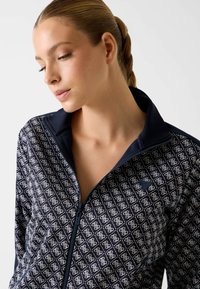 Granatowa kurtka na zamek z geometrycznym białym wzorem, teksturowaną tkaniną i wysokim kołnierzem. Widok z bliska na kołnierz i detale zamka.