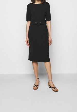 Frau trägt ein eng anliegendes schwarzes knielanges Kleid mit halblangen Ärmeln, einem breiten Gürtel und schwarze, ristrapazierte Sandalen mit niedrigem Absatz vor einfachem Hintergrund.