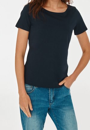 Vrouw in een donkerblauw T-shirt met korte mouwen en blauwe spijkerbroek, met één hand in de zak, tegen een witte achtergrond.