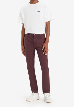Mann trägt weißes Levi’s T-Shirt, bordeauxfarbene Slim-Fit-Hose und weiße Sneaker, steht vor einfarbigem Hintergrund.