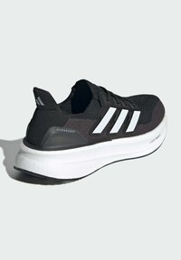 Svart träningsskor med stickad ovandel, tre vita ränder på sidan och en tjock vit sula märkt "Lightboost" och "Ultraboost."