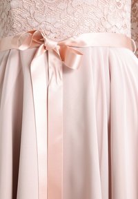 Spetsliv med ett lätt rosa satinband i midjan, som leder ner till en plisserad, flytande kjol i en matchande mjuk rosa färg.