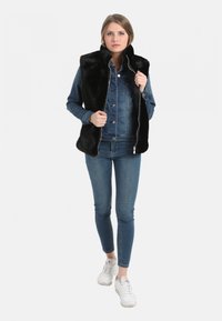 Gilet en fausse fourrure noire sur une chemise en denim bleu et un jean assorti. L'ensemble comprend des baskets blanches et présente une fermeture éclair à l'avant ainsi que des poches latérales.