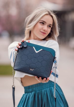 Modrá a černá kabelka s texturovaným povrchem, stříbrným kováním a odnímatelným řetízkem na rameno. Obsahuje logo a kontrastní horní panel.