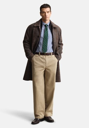 Soprabito a quadri marrone, camicia a righe blu e bianche, cravatta a righe verdi e bianche, pantaloni chino beige, scarpe marrone scuro, con le mani nelle tasche.