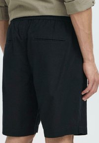 Stoffbukser i svart med en myk tekstur. Elastisk midjebånd med to baklommer. Rett snitt, som når opp til midten av låret. Minimalistisk design.