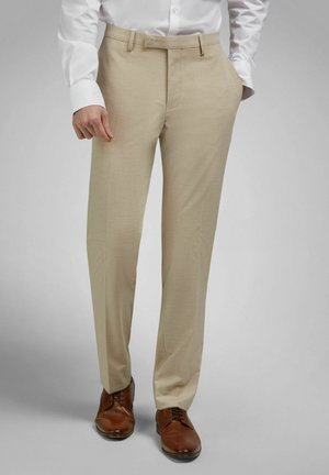 Mann trägt beige Stoffhose, weißes Hemd mit Knöpfen und braune Lederschuhe, steht mit der rechten Hand in der Tasche.