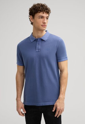 Junger Mann mit lockigem Haar, der ein blaues Polohemd mit kurzen Ärmeln und schwarze Hose trägt, leicht nach rechts blickend vor einem einfarbigen Hintergrund.