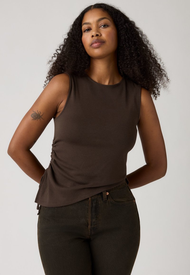 Femme aux cheveux bouclés portant un haut marron foncé sans manches et un jean foncé, posant avec les mains derrière le dos sur un fond uni.