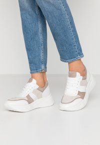 Personne portant des baskets en daim blanc et beige avec des semelles épaisses et un jean en denim bleu, debout sur un sol blanc.