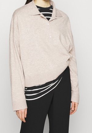Personne portant un pull beige à manches longues avec col par-dessus une chemise rayée noire et blanche, assorti d'un pantalon noir.
