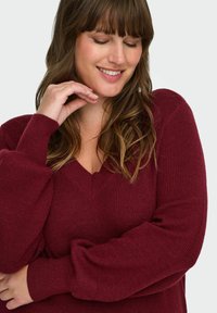 Maglione a maglia bordeaux con scollo a V, texture a coste e maniche a palloncino, caratterizzato da una vestibilità comoda e un materiale morbido.