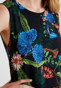 Débardeur noir sans manches orné de fleurs bleues vives, de feuilles vertes, de fleurs rouges et de papillons bleus sur une branche, porté par une personne.