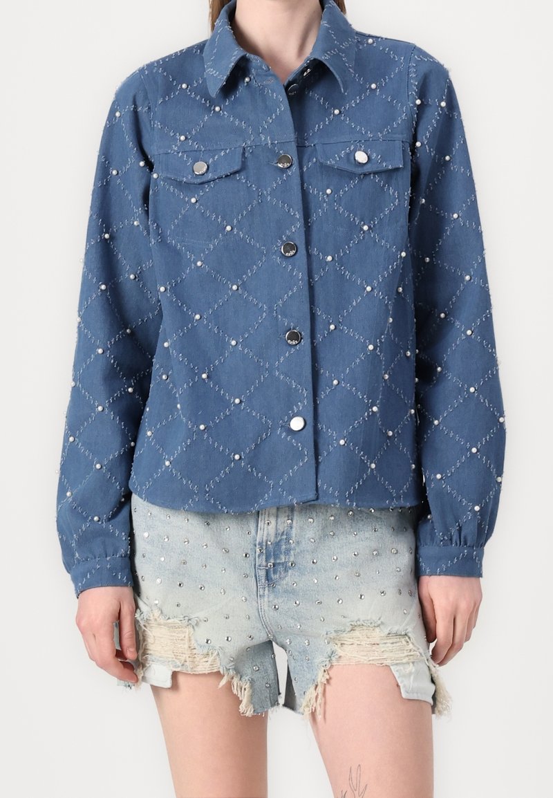 Veste matelassée bleue avec des clous argentés et des poches, associée à un short en jean clair déchiré avec des clous argentés.