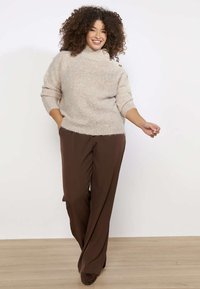 Žena s kučeravými vlasmi sa usmieva, má na sebe béžový rozopnutý sveter a voľné hnedé nohavice, stojí na drevenej podlahe pred bielou stenou.