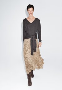 Frau mit kurzen dunklen Haaren, die einen dunklen V-Ausschnitt-Pullover trägt, der in der Taille gebunden ist, einen gemusterten beigen Rock und dunkelbraune Stiefel, stehend vor weißem Hintergrund.