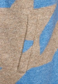 Ulltyg med en blandning av ljusbruna och blå färger, prytt med abstrakta former och en fickdetalj. Den strukturerade ytan är synlig.