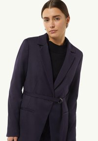 Granatowa marynarka z paskiem i klapami, posiadająca dwie przednie kieszenie oraz gładką teksturę. Noszona na czarnym swetrze.