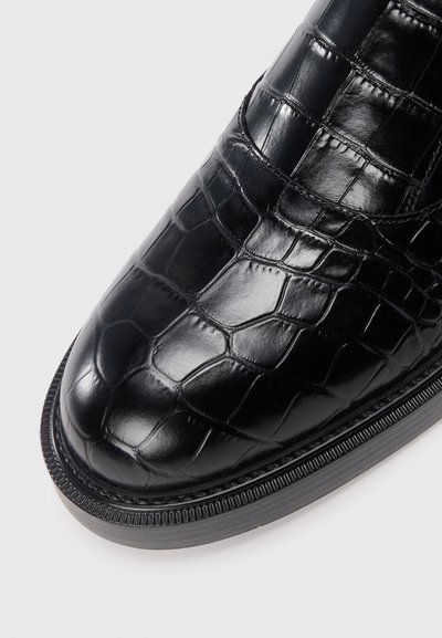 Schwarzer Lederschuh mit einem Krokodil-Muster, runder Spitze und strukturiertem Obermaterial. Die Sohle ist flach mit einem gerippten Design.