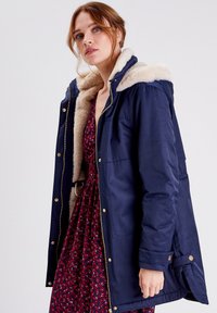Granatowa kurtka z kapturem wyłożonym sztucznym futrem, złotymi zamkami i guzikami, z zamkiem błyskawicznym oraz kieszeniami bocznymi. Miękka, gładka tekstura.