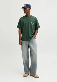 Grønn bomulls T-skjorte med korte ermer, med en liten hvit logo. Brukt med lyseblå baggy jeans og beige slip-on sko. Marineblå caps.